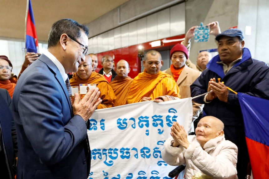 A crowd of Cambodian supporters stand behind a banner in Cambodian script and greet Sam Rainsy in an airport corridor.