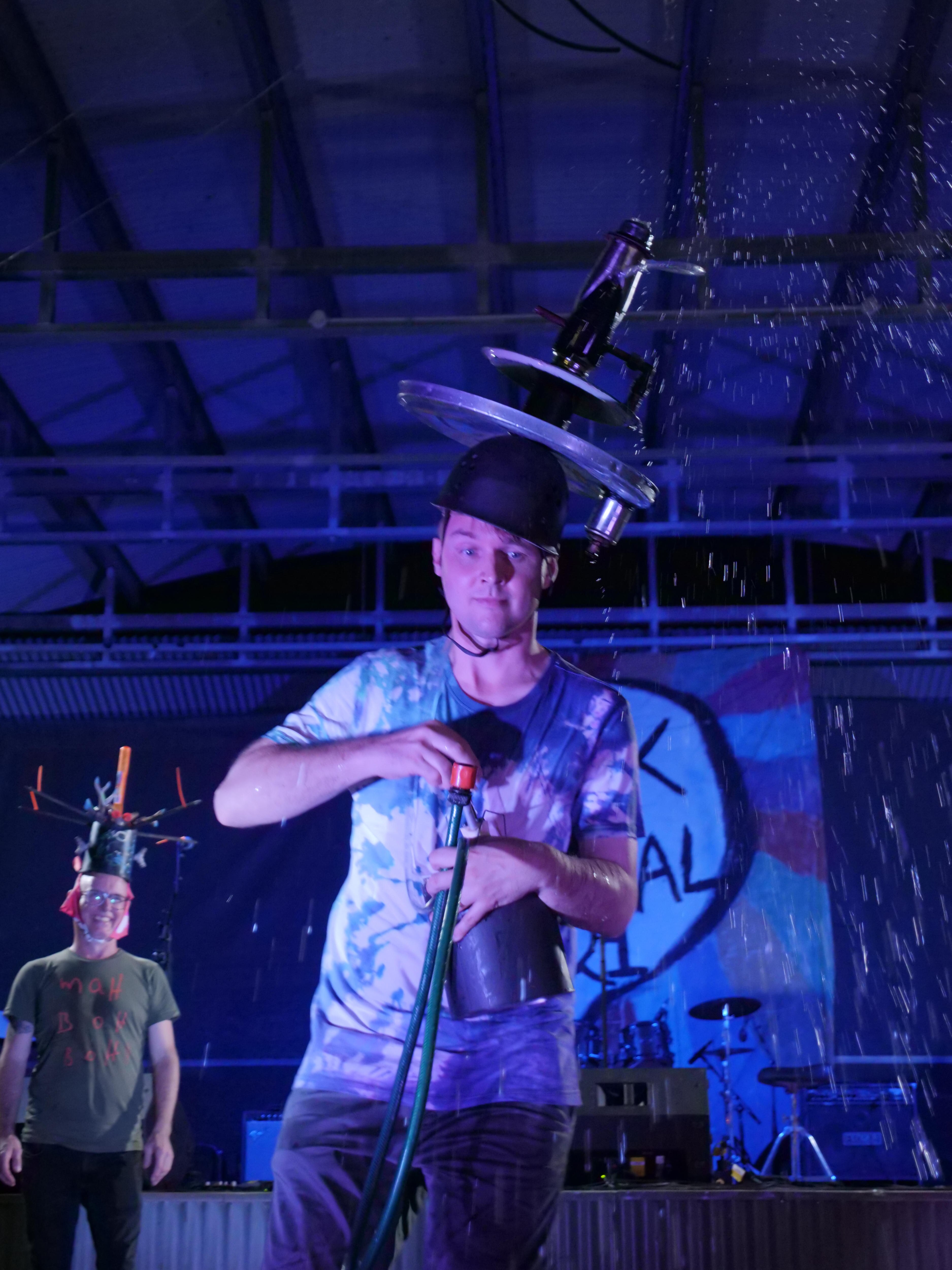 A man wearing a hat that spurts water in the air, using a pump. 