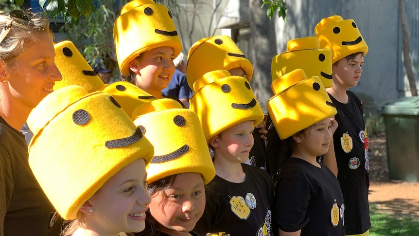 A group of school children with foam Lego man hats smiles while posing for a photo outside.