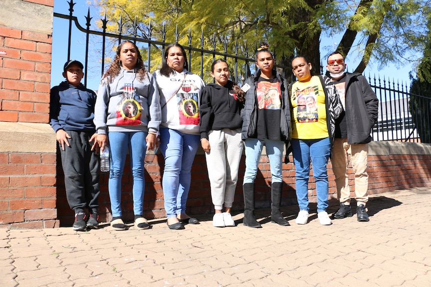 Seven people stand in front of a black metal bar fence with brick pillars