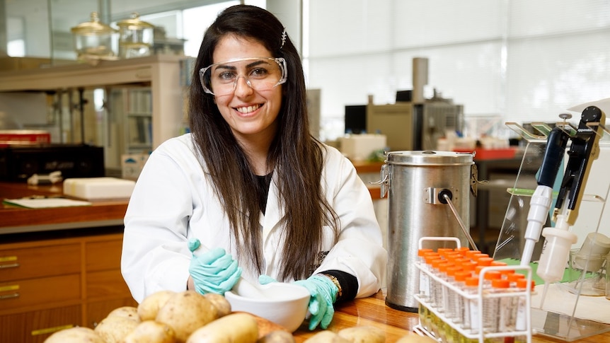 A female scientist with long, dark hair wearing safety glasses, sitting at a bench in front of a pile of potatoes.