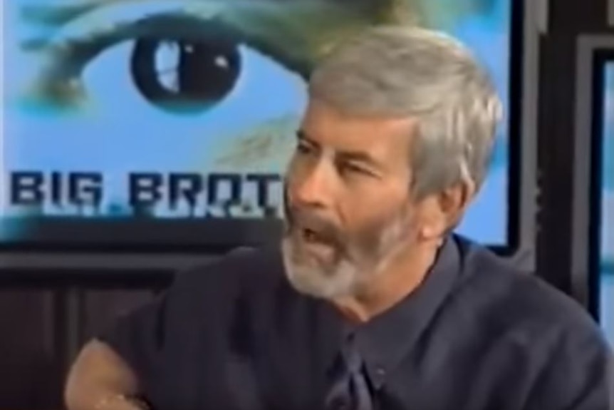A man with beard talking in front of a graphic of an eye with the "big brother" logo.