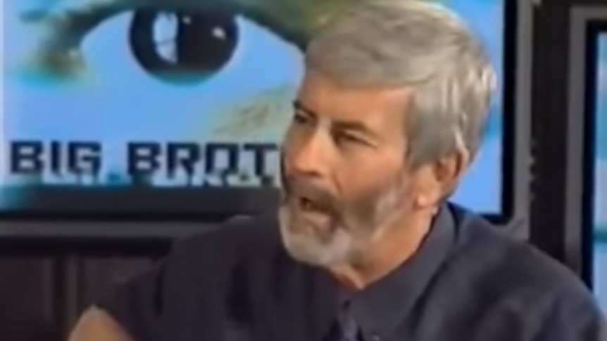 A man with beard talking in front of a graphic of an eye with the "big brother" logo.