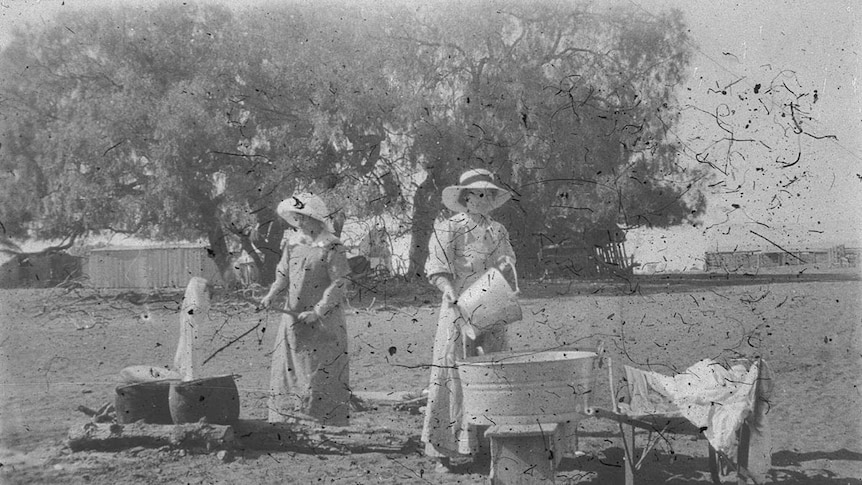 A black and white photo of women in 19th century attire doing laundry outside on an open fire with large peppercorn tree behind