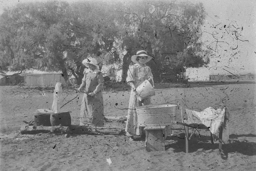 A black and white photo of women in 19th century attire doing laundry outside on an open fire with large peppercorn tree behind