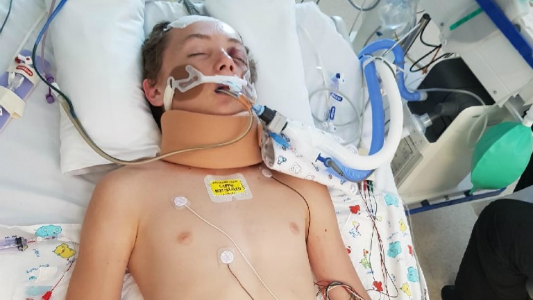 A teenage boy laying on a hospital bed with eyes closed and a lot of tubes attached to his body.