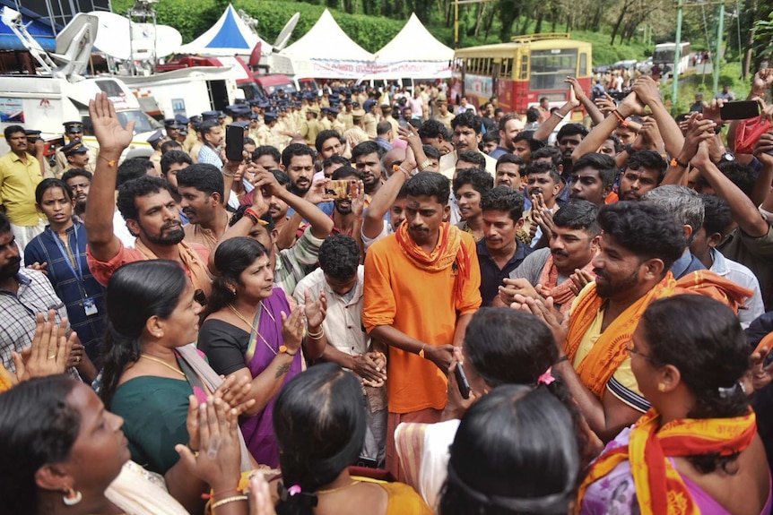 A group of Indian men and women in saris and scarfs chant in a circle as others film on their smartphones.