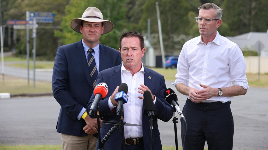 A male politician in a shirt and jacket speaking behind a microphone with two other men standing behind him.