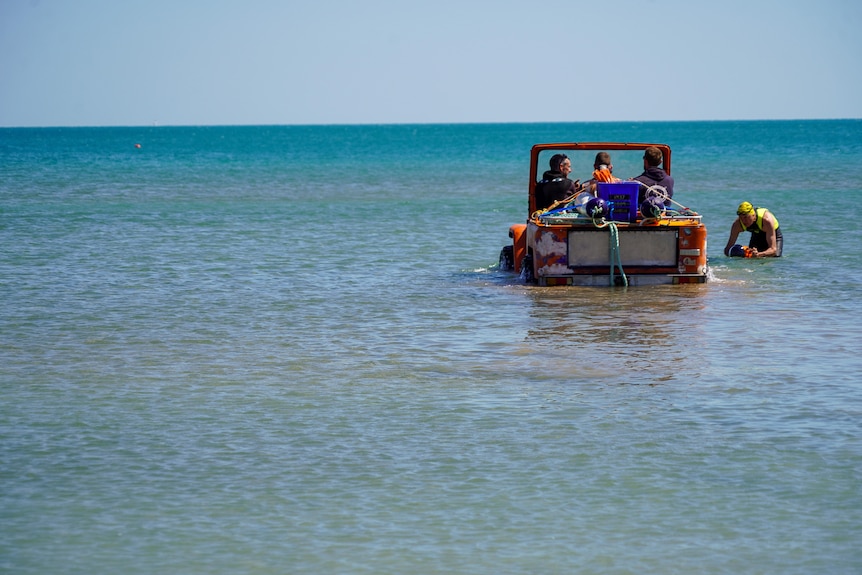 A car is being driven underwater with three men on board. The car is bright orange.