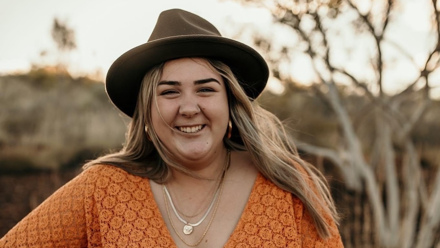 A young blonde woman wearing an orange dress and kahki hat smiles with country bush behind her