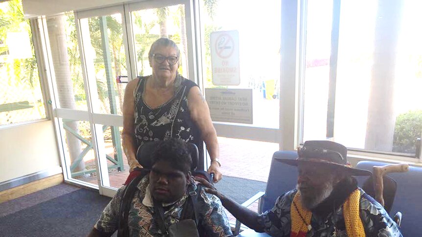 An older man comforts a teenager in a wheelchair as an older woman stands behind, at an airport lounge