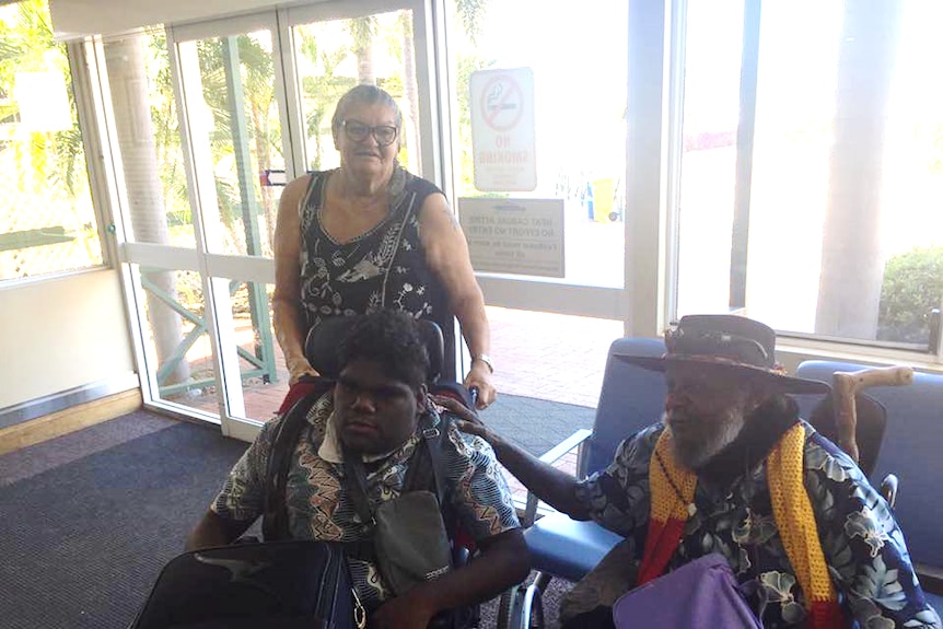 An older man comforts a teenager in a wheelchair as an older woman stands behind, at an airport lounge