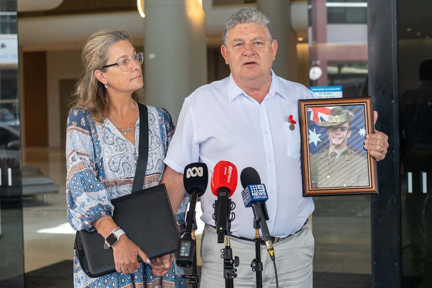 A middle-aged couple stands in front of reporters' microphones.  In his hand he holds a framed picture of a young man in military uniform