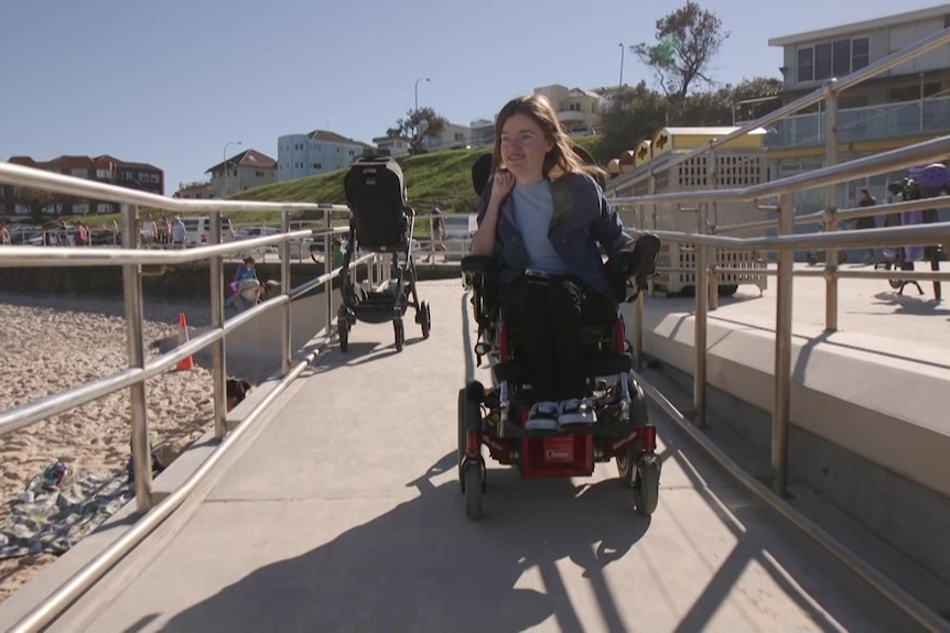 A woman in a motorised wheelchair moves down a ramp, towards the beach.