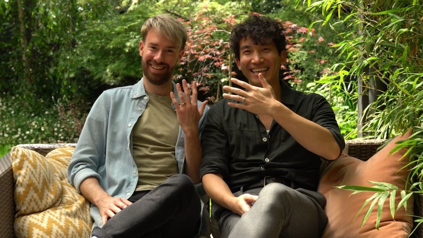 Two men sit on a lounge in a garden, holding up their hands to show their rings.
