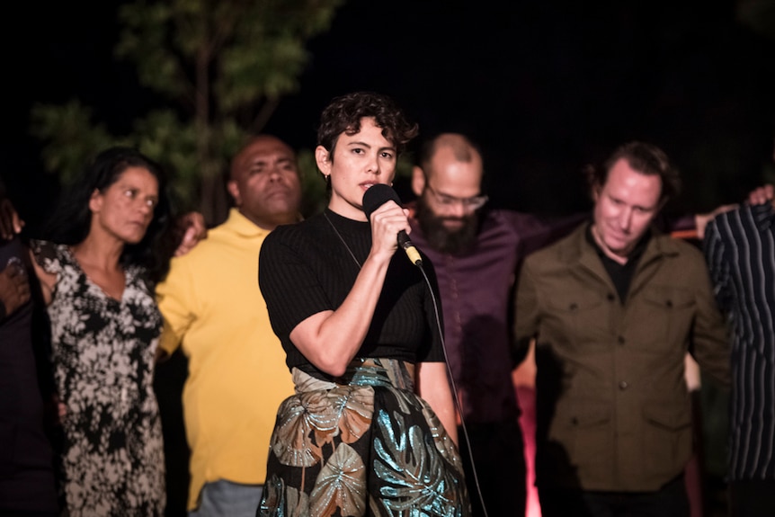 Exterior night-time show with woman with short wavy brown speaking into mic with tree and people behind her in a line.