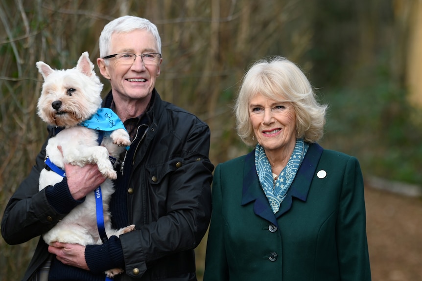 Paul O'Grady holds a small white dog while standing next to a smiling Camilla