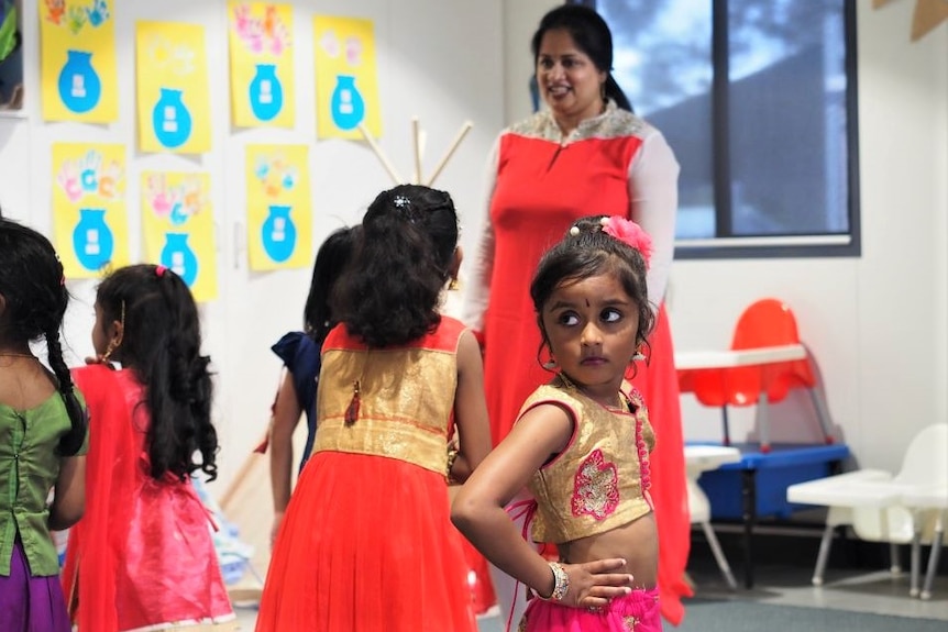 A young girl wearing a pink and gold sari stands with hands on hips. Her dance teacher and other students are in the background.