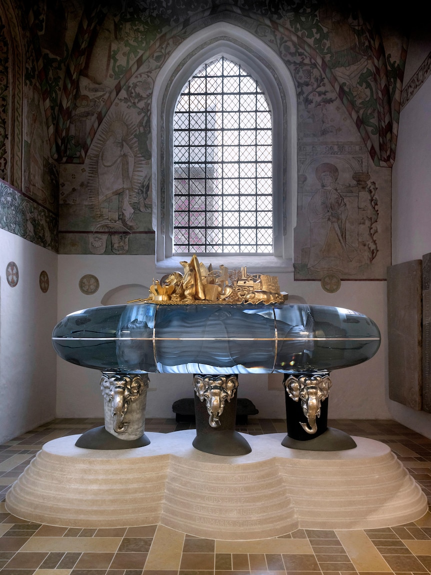 A large glass coffin is suspended on three silver elephant statues. On top sit gold adornments, a statue inside