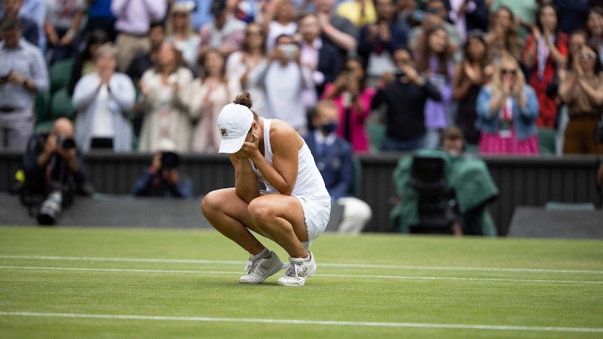 woman tennis player crouching with head in hands, on lawn tennis court, crowd standing and applauding behind her