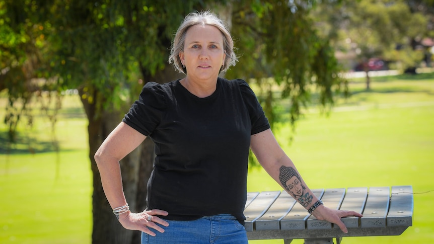 Serious woman, short blonde hair, tattoo of Lord Buddha on forearm, wears black tee, jeans, stands in park with wooden table.