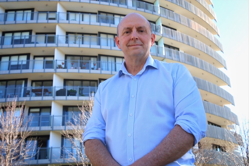 A man in a blue shirt stands in front of an apartment building.