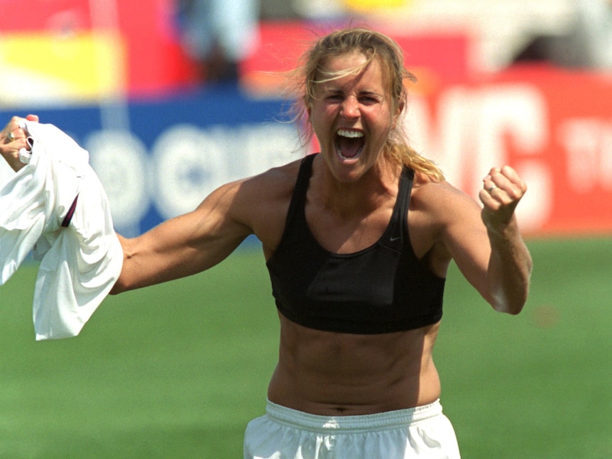 Brandi Chastain, wearing a crop top has her fists pumped and jersey in her hand.