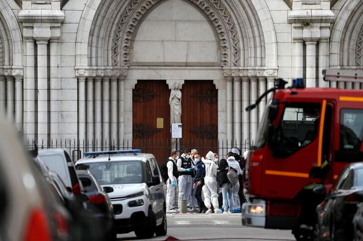 A grey stone church is seen with emergency vehicles in front of it and forensic workers outside the wooden doors.