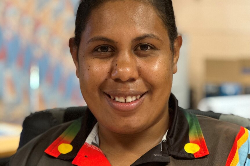 Image of a young woman smiling into the camera, she is wearing a polo with an Indigenous print.