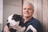a man smiles as he poses holding a black and white dog