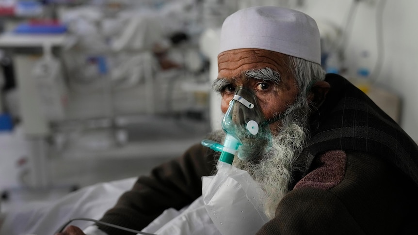 A man with a long white beard sits on a hospital bed, an oxygen mask over his nose and mouth.