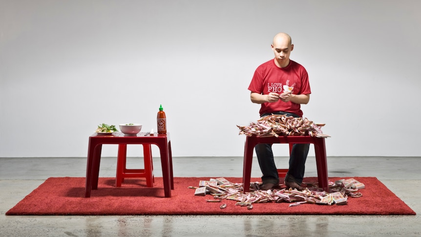 A man sits at a table making origami boats, next to a table of pho.