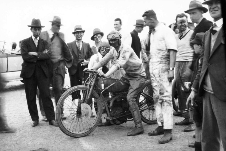 A man sits on a vintage motorcycle surrounded by people.