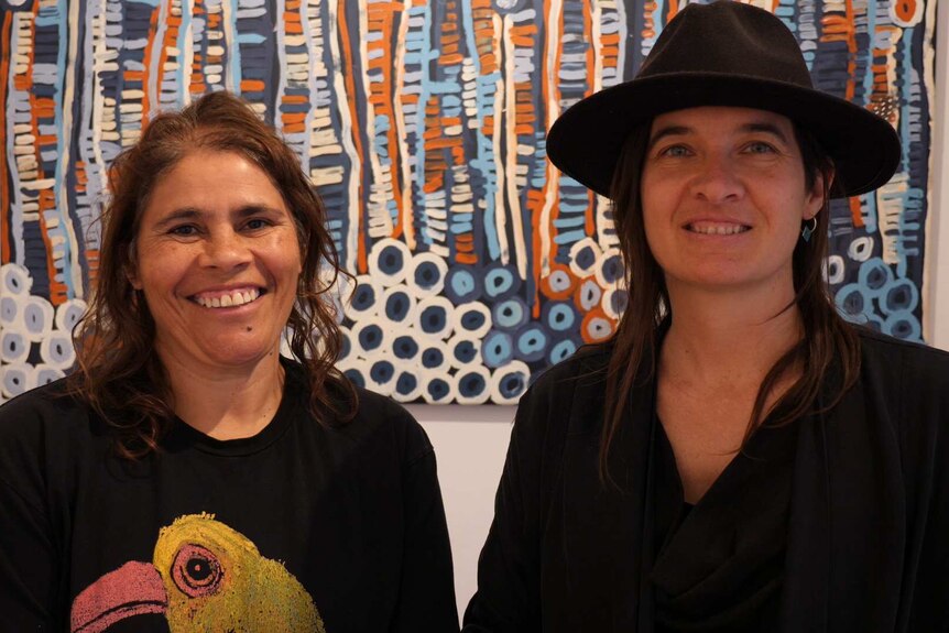 Two Aboriginal women wearing black and one with a hat are smiling in front of a painting.