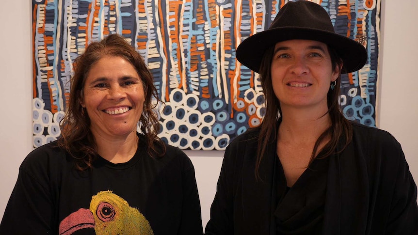 Two Aboriginal women wearing black and one with a hat are smiling in front of a painting.