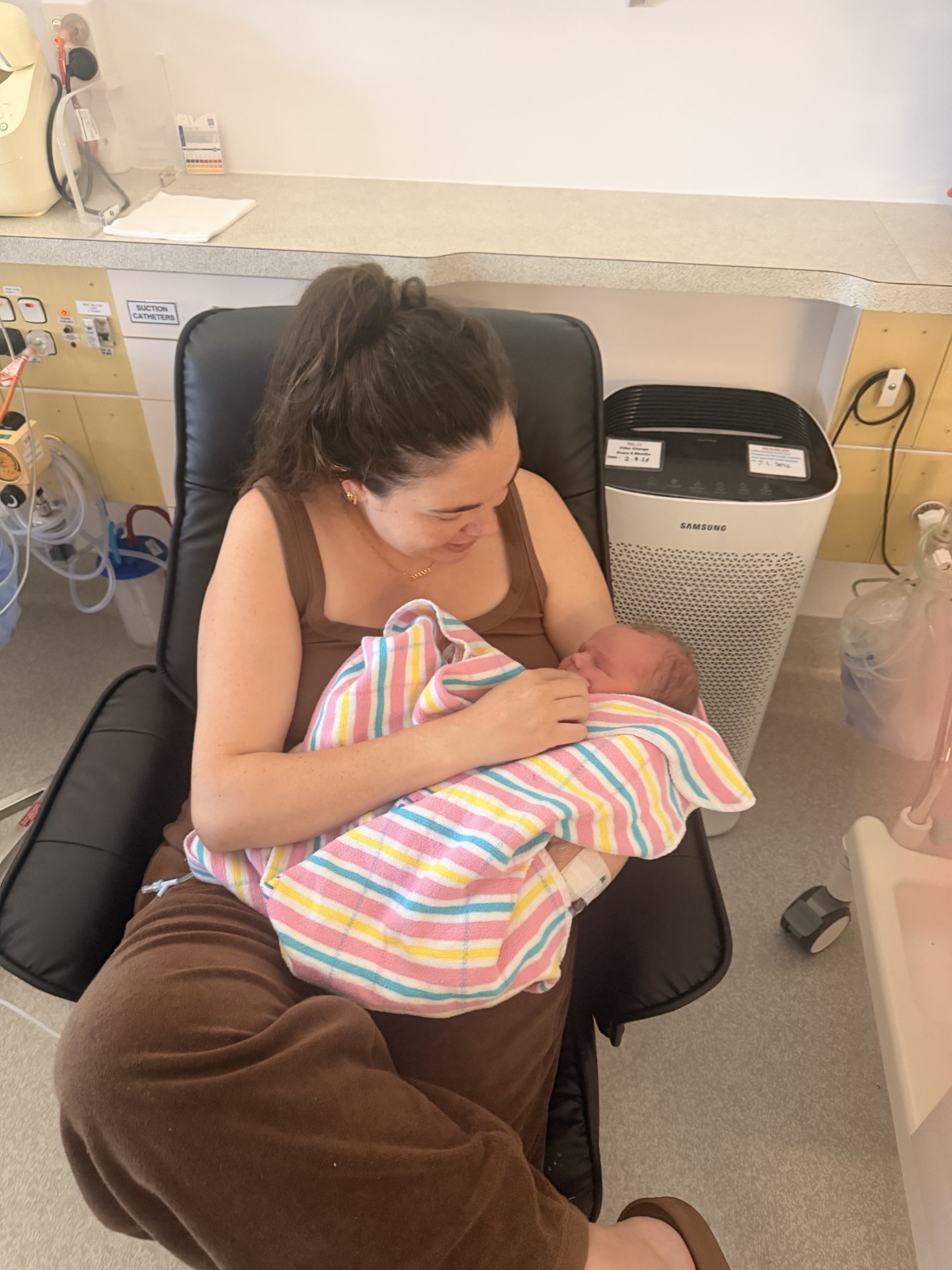 A woman in a brown dress nurses a newborn baby who is wrapped up in a blanket. 