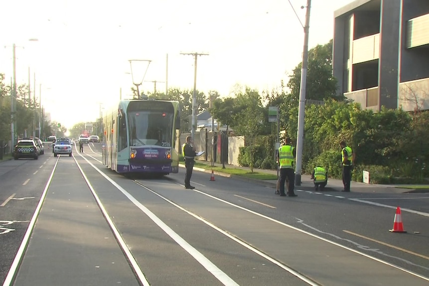 A tram stopped on its tracks with orange cones on the road as police stand around investigating the scene. 