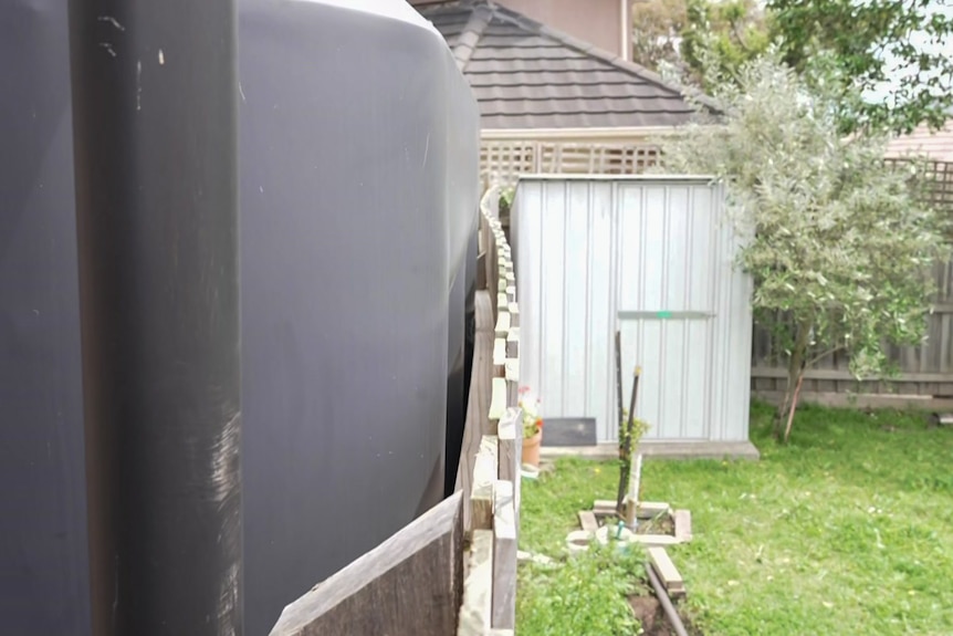 A large water tank sits right next to a wooden fence and is leaning on it, making the fence bulge out.