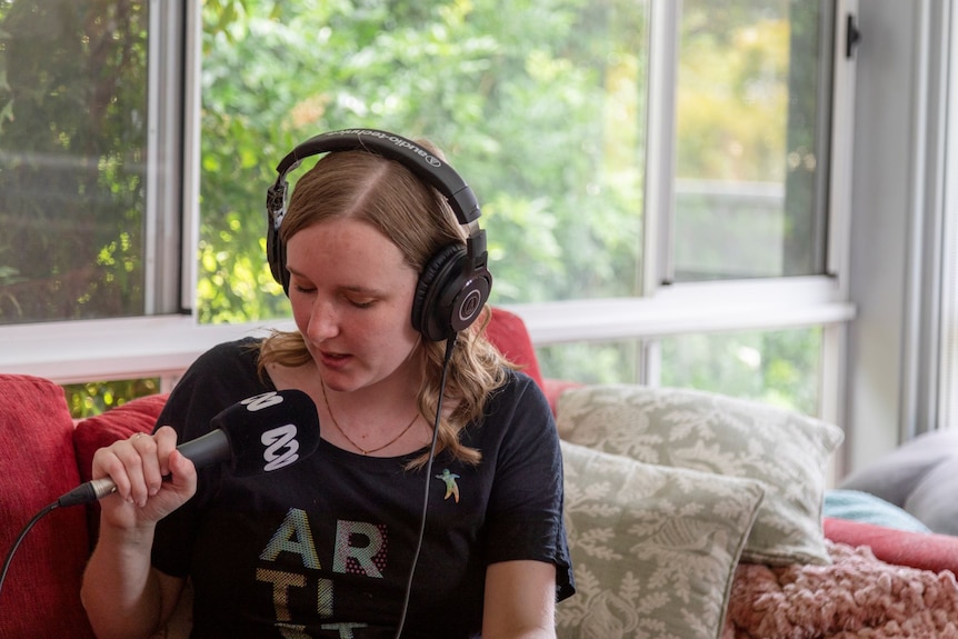 A woman with light brown hair sits on a couch holding an ABC microphone wearing headphones.