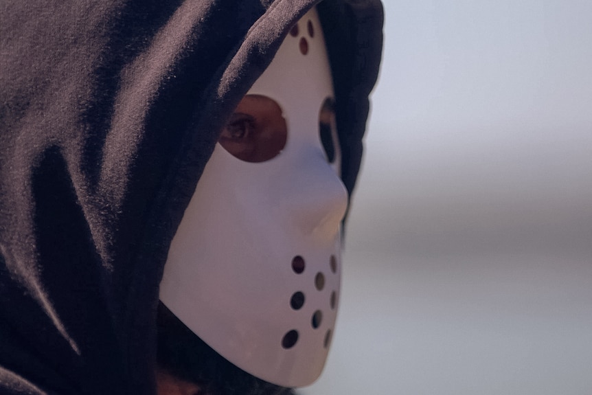 A man wearing a hoodie and a hockey mask, looks to the side, his face obscured.