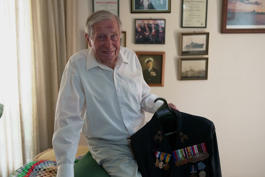 A man leans on a couch smiling, holding his jacket adorned with war medals