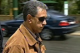 A side-on head and shoulders shot of Garry Narkle walking along a street wearing a brown jacket and sunglasses.