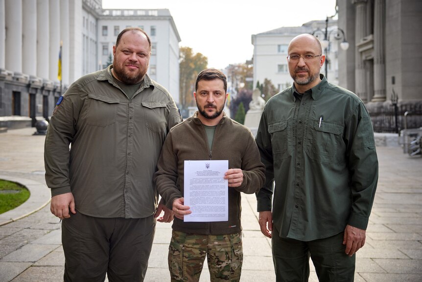 Ukraine's president holds up a piece of paper, flanked by two men either side.