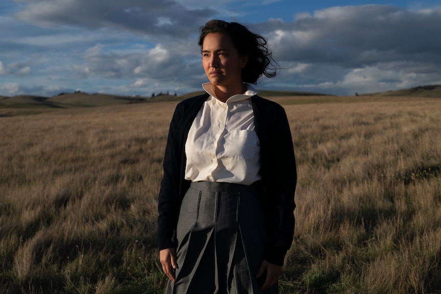 A woman standing in a field, serious expression, wearing a cardigan, white shirt and skirt, blue sky behind