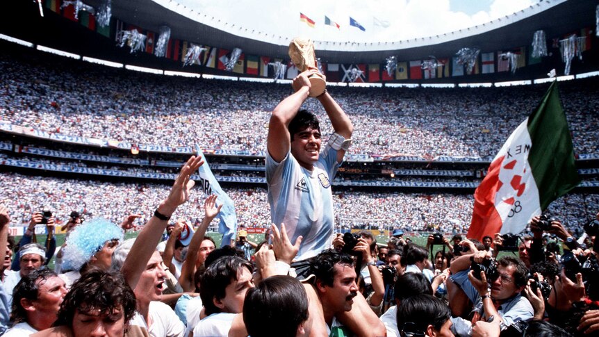Maradona is hoisted up by his team mates, holding the gold FIFA trophy above his head, with a stadium full of people looking on.