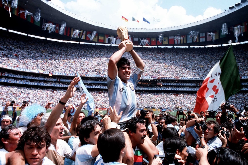 Maradona is hoisted up by his teammates, holding the gold FIFA trophy above his head, with a stadium full of people looking on.
