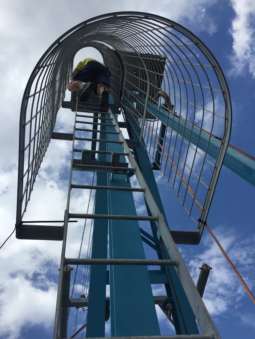 A man in a flouro yellow work shirt and blue pants climbing up a ladder to the highest platform on a crane.