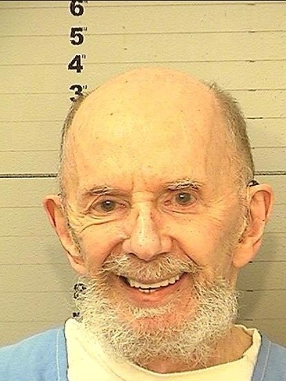 An elderly bald man with a goatee smiles as he poses for a headshot while in custody.
