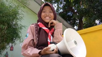 A young girl wearing a head scarf holds a speakerphone and smiles