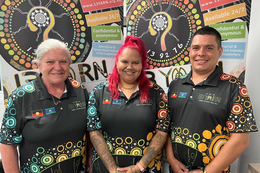 Three employees of 13YARN, wearing shirts with Indigenous designs, smile. Two ladies and one man.